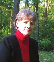 A woman with short gray hair wearing a red turtleneck and black sweater stands in front of a forested background.